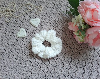 May include: White crocheted hair scrunchie on a gray crocheted surface with two white ceramic hearts and a bouquet of pink peonies.
