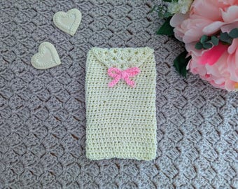 Puede incluir: Una funda de teléfono de ganchillo hecha a mano, de color amarillo claro, con un lazo rosa. La funda rectangular tiene un borde superior festoneado. También se ven dos decoraciones blancas en forma de corazón y peonías rosas.