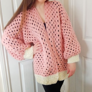 May include: A pink crocheted cardigan with white trim on the cuffs and hem. The cardigan has a loose fit and a V-neck.