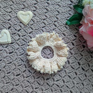 May include: A close-up of a crocheted hair scrunchie in a light cream color. The scrunchie is lying on a gray crocheted surface. Two white heart-shaped decorations are also visible on the surface.