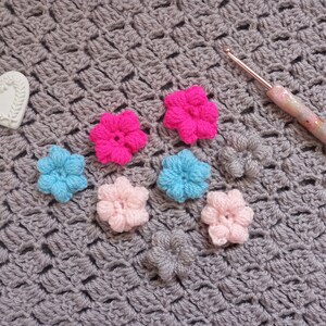 May include: A close-up of a gray crocheted blanket with several crocheted flowers in pink, blue, and hot pink. A white heart-shaped ornament and a pink and white crochet hook are also visible.