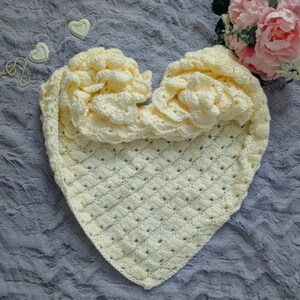 May include: A cream-colored, heart-shaped crocheted blanket with a textured pattern. The blanket is folded to display a ruffled edge. Decorative elements include a string of pearls, two heart-shaped ornaments, and a bouquet of pink flowers.