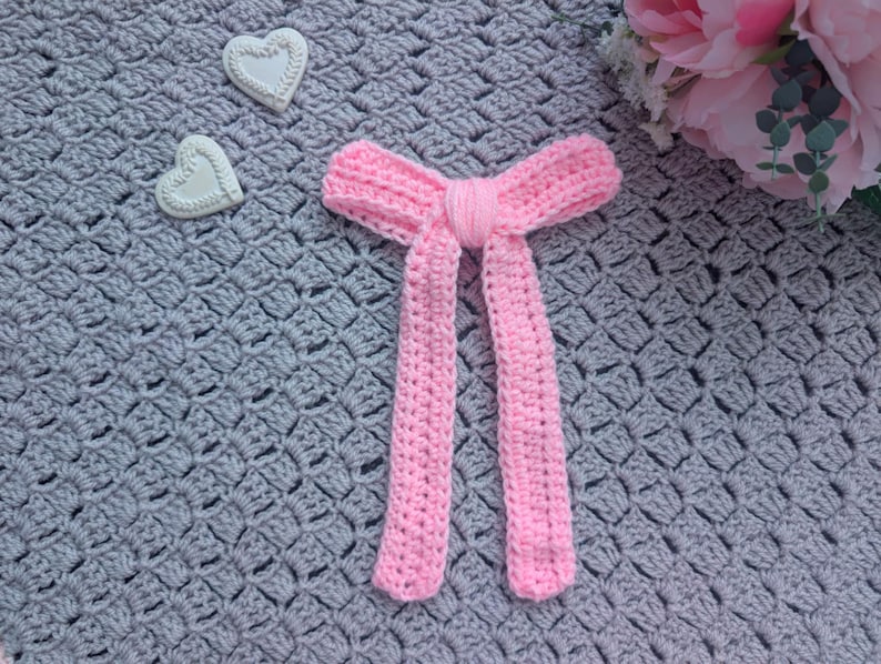 Peut inclure: Un n&oelig;ud papillon rose en crochet fait &agrave; la main, avec de longues extr&eacute;mit&eacute;s pendantes. Le n&oelig;ud est centr&eacute; sur une couverture grise en crochet, avec deux d&eacute;corations en forme de c&oelig;ur blanc et des fleurs roses en arri&egrave;re-plan.