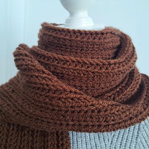 May include: A brown crocheted scarf with a textured pattern. The scarf is folded and draped over a white mannequin.