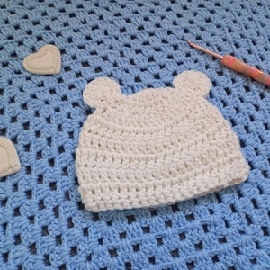 May include: A white crocheted baby hat with bear ears on a blue crocheted blanket. A pink crochet hook is also in the image.