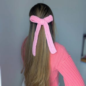 Peut inclure: Un accessoire pour cheveux en forme de n&oelig;ud rose clair, crochet&eacute;. Le n&oelig;ud est nou&eacute; au centre avec de longues extr&eacute;mit&eacute;s pendantes. L'accessoire pour cheveux est port&eacute; dans les cheveux. La personne porte un pull rose.