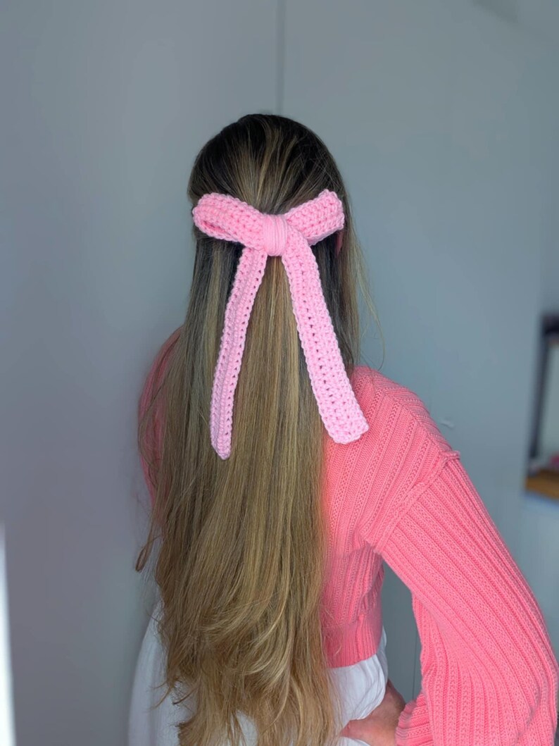 Peut inclure: Un accessoire pour cheveux en forme de n&oelig;ud rose clair, crochet&eacute;. Le n&oelig;ud est nou&eacute; au centre avec de longues extr&eacute;mit&eacute;s pendantes. L'accessoire pour cheveux est port&eacute; dans les cheveux. La personne porte un pull rose.