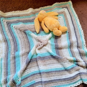 Może przedstawiać: Kocyk dla niemowląt wykonany na szydełku w pasy w kolorze białym, beżowym i niebieskim. Kocyk ma haftowany brzeg i jest złożony na pół z pluszową zabawką na górze.