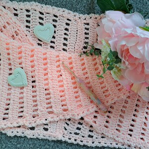 May include: A pink crocheted blanket with a delicate openwork pattern. Two white heart-shaped decorations are placed on the blanket. A crochet hook is also visible.
