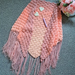 May include: A crocheted shawl with a gradient of pink and peach colours. The shawl has a fringe border and is laid flat on a grey carpet. Two small white hearts and a crochet hook are also visible in the image.