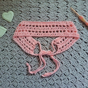 May include: A pink crocheted headband with two long ties. The headband is laid out on a grey crocheted surface.