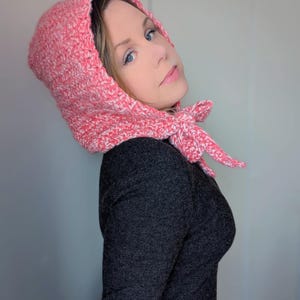 Peut inclure: Une cagoule rose et blanche en crochet avec une fermeture à nouer. La cagoule est faite d'un fil épais et est portée sur un haut à manches longues gris foncé. Un accessoire douillet.