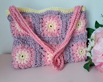 May include: A crocheted handbag with a pink and purple floral design. The bag has a long strap and is made with a textured yarn.