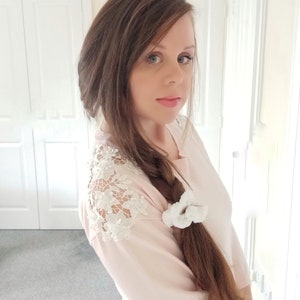 May include: A woman with long brown hair wearing a pink sweater with white lace detailing on the shoulder. She has a white scrunchie in her hair.
