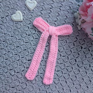 Peut inclure: Un n&oelig;ud papillon rose clair fait &agrave; la main au crochet, avec de longues extr&eacute;mit&eacute;s pendantes. Il est pos&eacute; sur une couverture grise au crochet, avec deux d&eacute;corations en forme de c&oelig;ur blanches et un bouquet de fleurs roses en arri&egrave;re-plan.