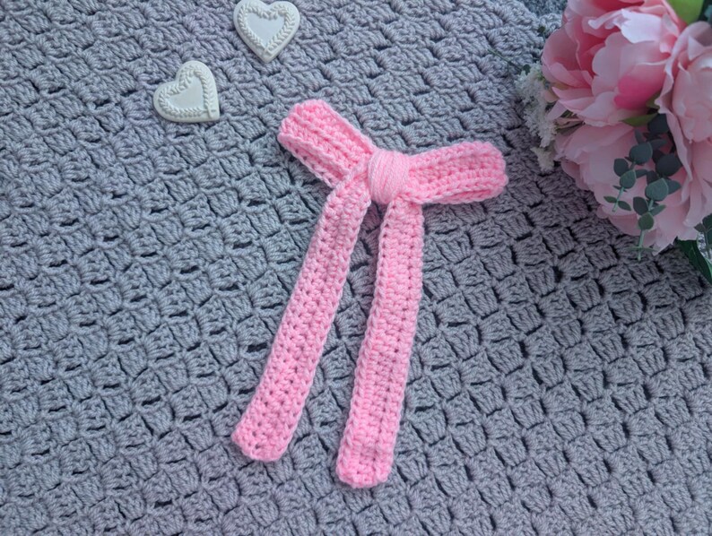 Peut inclure: Un n&oelig;ud papillon rose clair fait &agrave; la main au crochet, avec de longues extr&eacute;mit&eacute;s pendantes. Il est pos&eacute; sur une couverture grise au crochet, avec deux d&eacute;corations en forme de c&oelig;ur blanches et un bouquet de fleurs roses en arri&egrave;re-plan.