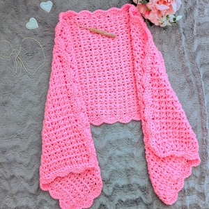 Peut inclure: Un boléro rose vif en crochet, avec de larges manches évasées et une bordure festonnée. Un crochet repose sur le vêtement. Le boléro fait main est présenté sur une surface grise avec des éléments décoratifs.
