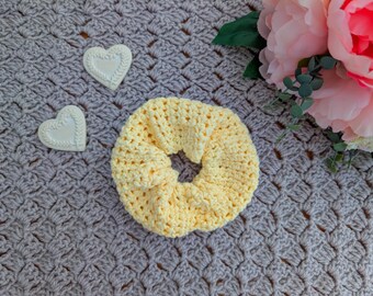 May include: A handmade yellow crochet scrunchie sits on a gray crocheted surface, next to two white heart-shaped decorations. Pink and white flowers with green leaves are in the background. The scrunchie is a soft, circular hair accessory.