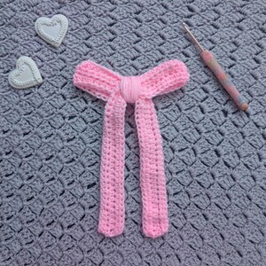Peut inclure: Un n&oelig;ud papillon rose en crochet avec de longues extr&eacute;mit&eacute;s pendantes. Deux d&eacute;corations en forme de c&oelig;ur blanc et un crochet avec un manche color&eacute; sont &eacute;galement visibles. L'ensemble est pos&eacute; sur une surface grise textur&eacute;e.