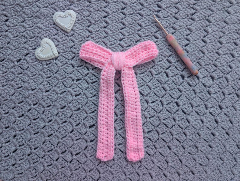 Peut inclure: Un n&oelig;ud papillon rose en crochet avec de longues extr&eacute;mit&eacute;s pendantes. Deux d&eacute;corations en forme de c&oelig;ur blanc et un crochet avec un manche color&eacute; sont &eacute;galement visibles. L'ensemble est pos&eacute; sur une surface grise textur&eacute;e.