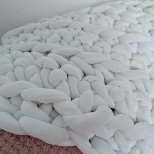 May include: A close-up of a white, chunky knit blanket. The blanket is made of thick, soft material, with a braided design. The white color gives it a clean and modern look. The blanket is a cozy home decor item.