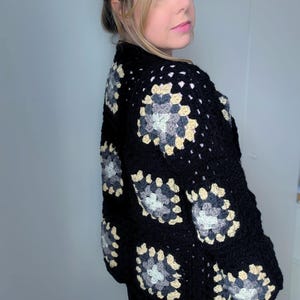 Puede incluir: Cárdigan negro de crochet con diseño de cuadrados de la abuela. El cárdigan presenta un patrón repetido de cuadrados negros, grises y beige. Una prenda hecha a mano y acogedora, perfecta para combinar.