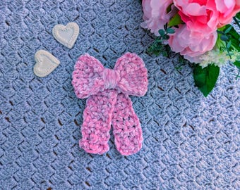 Puede incluir: Una pajarita rosa hecha a mano de ganchillo con una textura suave y aterciopelada, sobre una manta azul de ganchillo. Dos decoraciones blancas en forma de corazón están a la izquierda de la pajarita. Flores rosas y blancas en la esquina superior derecha.