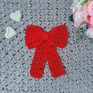 May include: A red crocheted bow with a lacy design. The bow is on a grey crocheted surface.