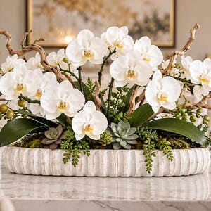 May include: A floral arrangement featuring white phalaenopsis orchids, green succulents, and moss in a long, white, ribbed oval planter. The display includes curved, light brown decorative branches and small green berries.