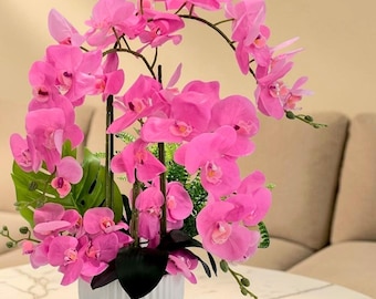 May include: A vibrant arrangement of pink orchids in a white, ribbed ceramic vase. The orchids are in full bloom, with lush green leaves and stems. The arrangement is set on a marble-topped table, with a neutral background.