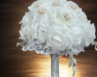 May include: A white bridal bouquet with pearl accents and a silver glitter stem. The bouquet is made of silk roses and lace.