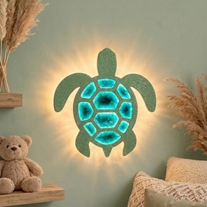 May include: A wall-mounted sea turtle light fixture featuring a glittery green outer shell and a glowing turquoise centre. The light emits a warm, soft radiance from behind the turtle shape, illuminating the surrounding area with a gentle, ambient glow.