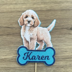 May include: A dog-themed cake topper featuring a light brown and white dog illustration. The dog stands above a blue bone-shaped sign with the name "Karen". The sign has a glittery texture.