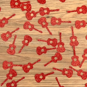 May include: Red glitter guitar-shaped confetti scattered on a light brown wooden surface. The confetti pieces are uniformly red with a sparkling texture. The image is a top-down view, showcasing the decorative elements.
