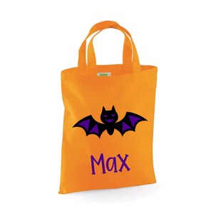 Puede incluir: Bolsa naranja de truco o trato de Halloween con un murci&eacute;lago negro y alas moradas. La bolsa tiene el nombre "Max" impreso en ella.