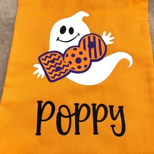 Puede incluir: Bolsa naranja de truco o trato de Halloween con un fantasma blanco que sostiene un dise&ntilde;o de caramelo morado y naranja. La bolsa est&aacute; personalizada con el nombre "Poppy".
