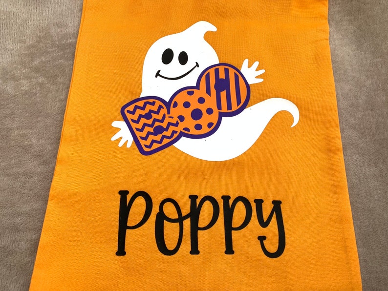 Puede incluir: Bolsa naranja de truco o trato de Halloween con un fantasma blanco que sostiene un dise&ntilde;o de caramelo morado y naranja. La bolsa est&aacute; personalizada con el nombre "Poppy".