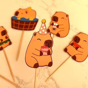 May include: A selection of charming capybara-themed cupcake toppers. Each topper depicts a cartoon capybara in different poses, including one with a birthday cake, one with a drink, and one with watermelon. The toppers are attached to wooden sticks.