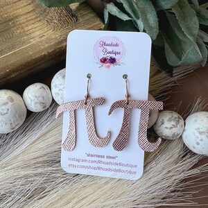 May include: Rose gold-colored earrings in the shape of the mathematical symbol pi, hanging from gold-colored hooks. The earrings are displayed on a white card with the text "Rhoadside Boutique". The earrings are made of stainless steel.