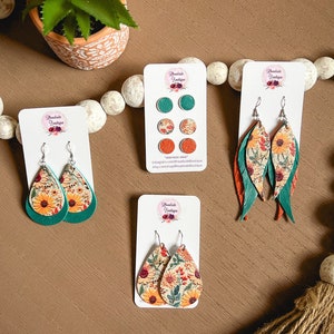 May include: Three pairs of earrings with floral patterns on a brown wooden surface. The earrings are made of leather and have a teal and orange color scheme. The earrings are displayed on white cards with the text "Remarkable Boutique" on them.