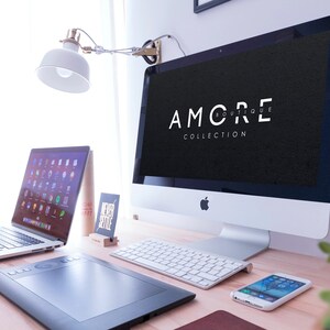 May include: A white desktop computer with a black screen displaying the text "AMORE BOUTIQUE COLLECTION". The computer is on a wooden desk with a laptop, a wireless keyboard, a drawing tablet, and a smartphone.
