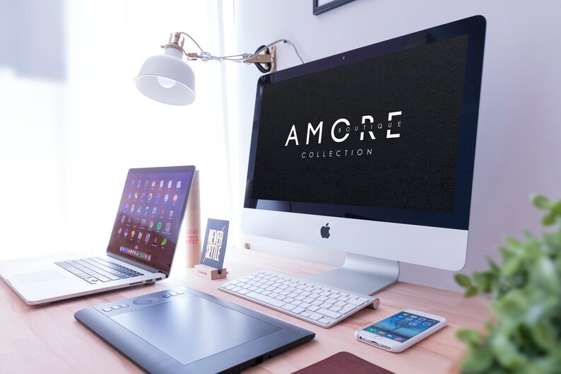 May include: A white desktop computer with a black screen displaying the text "AMORE BOUTIQUE COLLECTION". The computer is on a wooden desk with a laptop, a wireless keyboard, a drawing tablet, and a smartphone.