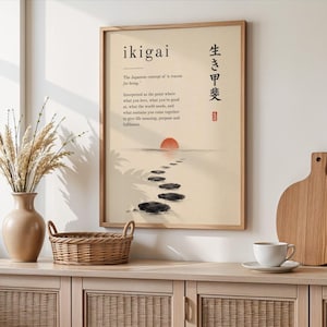 May include: A framed wall art print featuring the word ikigai, Japanese characters, and a definition text. The illustration depicts a red sun over a path of dark stones leading into the distance on a cream background, presented in a light wood frame.