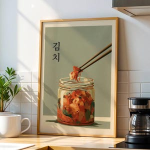 Könnte beinhalten: Ein gerahmter Kunstdruck mit einem Glas Kimchi, wobei Essstäbchen einen Teil des Essens halten. Der Druck hat einen hellgrünen Hintergrund mit dem koreanischen Wort für Kimchi. Der Druck ist in einem Holzrahmen.