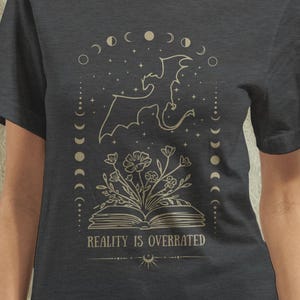 May include: Dark grey t-shirt with a celestial design. It features a dragon, moon phases, and flowers emerging from an open book. The text "REALITY IS OVERRATED" is printed below the book. The design is in a light tan colour.
