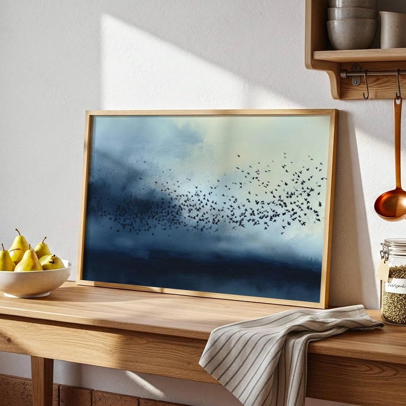 May include: A rectangular framed wall art print featuring a dark blue and cream gradient background with a large flock of small black birds in flight. The artwork is displayed on a wooden surface next to a bowl of yellow pears and a striped cloth.