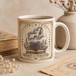 Könnte beinhalten: Weiße Keramiktasse mit Fantasy-Design. Die Tasse zeigt einen Drachen, Blumen und den Text "REALITY IS OVERRATED" im Vintage-Stil. Die Tasse ist mit einer dunklen Flüssigkeit gefüllt.