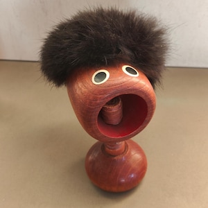 Pode incluir: Um quebra-nozes de madeira com um chapéu de pele marrom e grandes olhos pretos. O quebra-nozes é vermelho e marrom e tem uma boca que se abre para quebrar nozes.