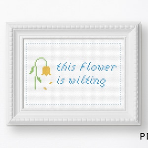 May include: A white framed cross-stitch with blue and yellow thread. The cross-stitch depicts a wilting yellow flower with the text "This flower is wilting".