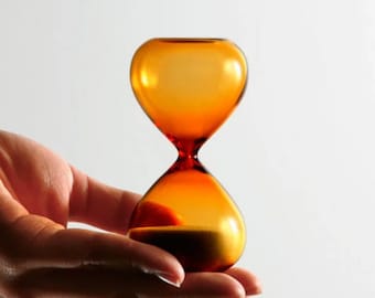 May include: An amber glass hourglass with sand flowing through the center. The hourglass is being held in a person's hand.
